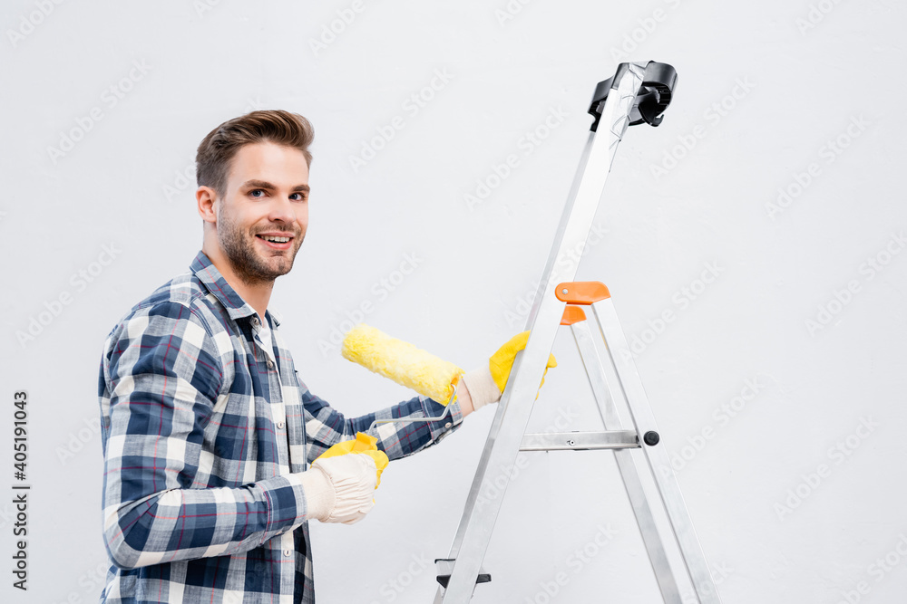 happy young man looking at camera while holding paint roller on ladder isolated on white happy young man looking at camera while holding paint roller on ladder isolated on white