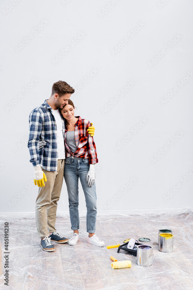Fototapeta premium full length of smiling young couple hugging near tins of paint, rollers and tray at home