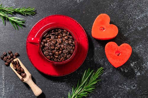 Festive cafeteria or coffee shop banner with red cup full of coffee beans, heart-shaped candles and aromatic rosemary twigs on black rustic background top view. St. Valentine’s Day concept.