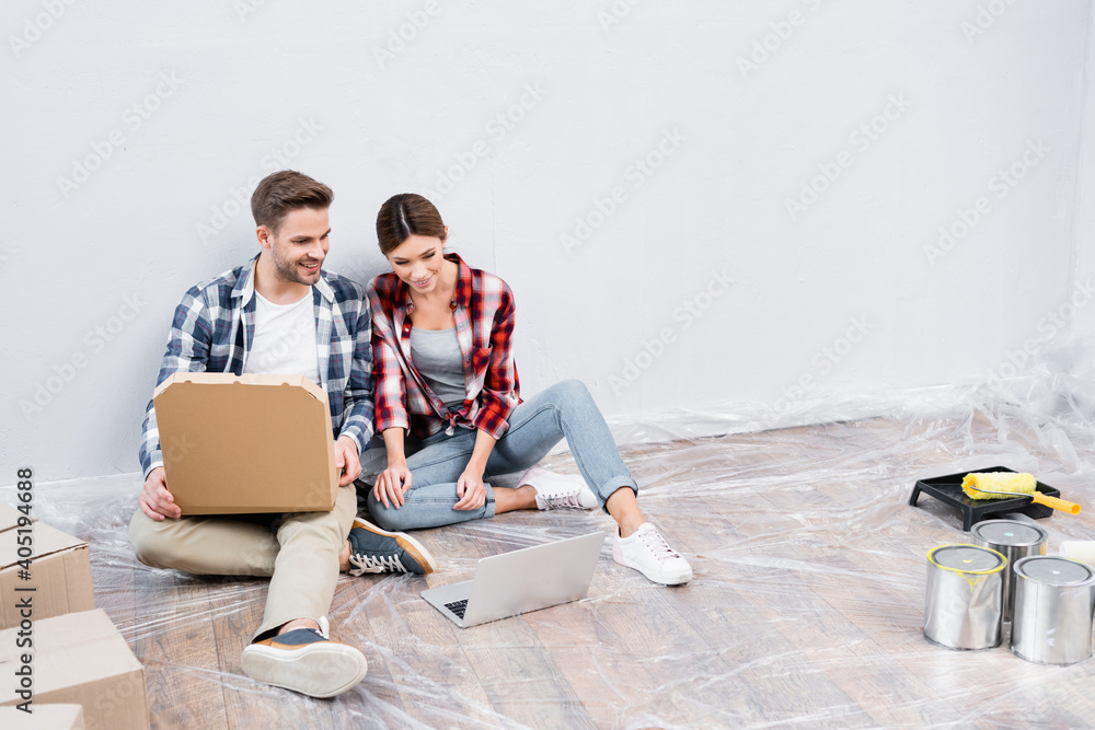 full length of smiling young man with cardboard box of pizza watching film on laptop near woman while sitting on floor at home