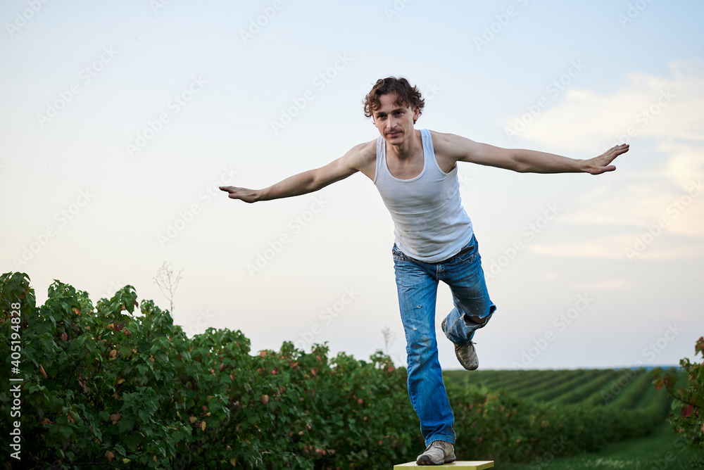 Young handsome man, wearing torn jeans and white t-shirt, sitting on yellow stool on green field, thinking. Artistic education concept. Creative portrait outside. Young handsome man, wearing torn jeans and white t-shirt, sitting on yellow stool on green field, thinking. Artistic education concept. Creative portrait outside.