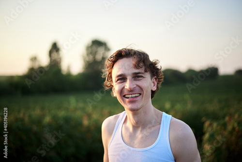 Young handsome man, wearing white t-shirt, smiling, laughing in front of green field. Human emotions concept. Happiness. Close-up male portrait outside on natural background.