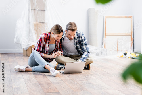 happy young couple looking at laptop while sitting on floor on blurred foreground at home