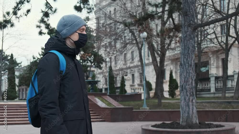 A man in black medical mask and a winter jacket with blue backpack looking into camera, building and park on the background in Chisinau, Moldova A man in black medical mask and a winter jacket with blue backpack looking into camera, building and park on the background in Chisinau, Moldova