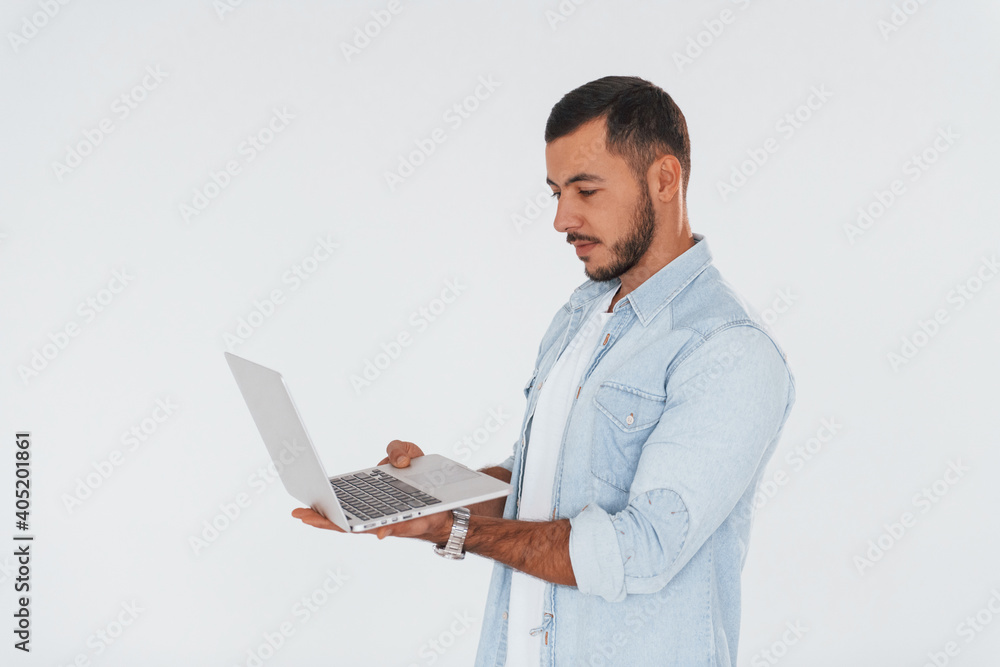 Uses laptop. Young handsome man standing indoors against white background Uses laptop. Young handsome man standing indoors against white background