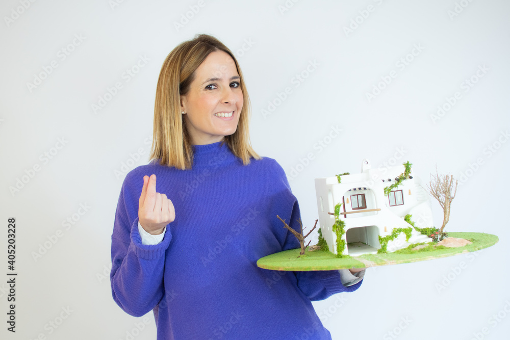 young pretty woman smiling cheerfully, feeling happy and showing a concept in copy space with palm of hand with a house model young pretty woman smiling cheerfully, feeling happy and showing a concept in copy space with palm of hand with a house model
