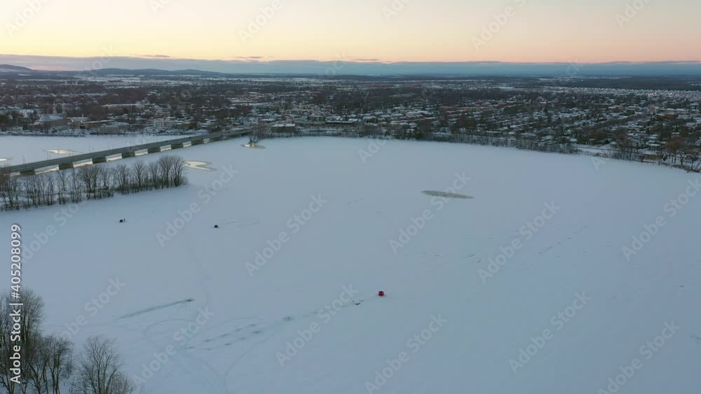 High aerial drone shot coming down slowly revealing the surrounding city and the ice fishing hut on the frozen river with some fishermen at sunset High aerial drone shot coming down slowly revealing the surrounding city and the ice fishing hut on the frozen river with some fishermen at sunset