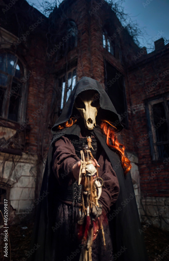 Scary red-haired witch with a staff in a black hooded cloak and a skull at night against the background of an old sinister castle Scary red-haired witch with a staff in a black hooded cloak and a skull at night against the background of an old sinister castle