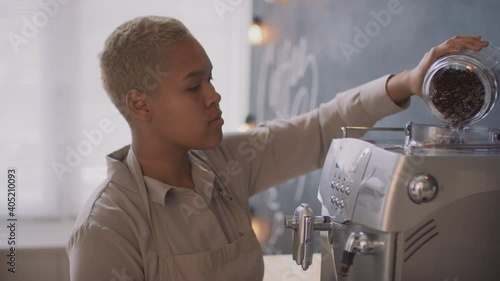 Side-view medium close-up footage of young happy black woman working in small coffee house indoors pouring fresh coffee beans into electric coffee machine, preparing cafe for opening photo