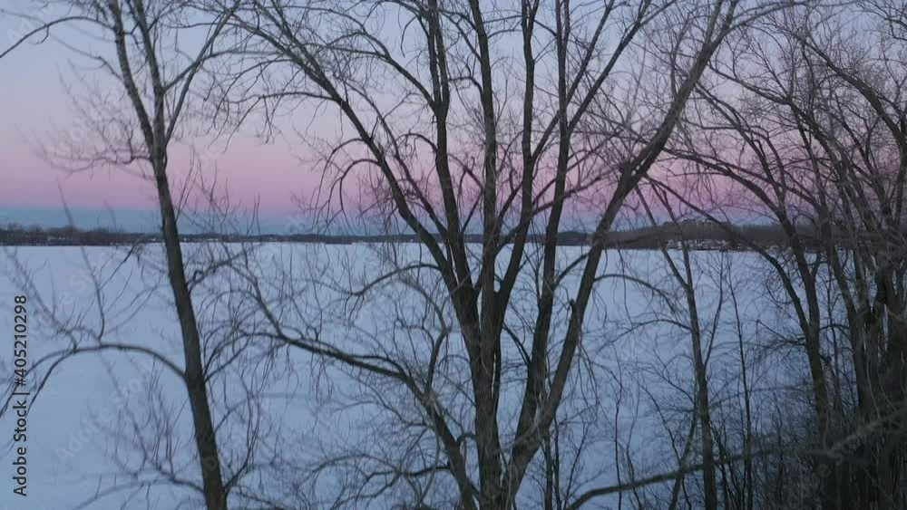 Drone shot rising from behind the trees and passing above them revealing the sunset with pink and blue colors and the fronzen river covered of white snow Drone shot rising from behind the trees and passing above them revealing the sunset with pink and blue colors and the fronzen river covered of white snow