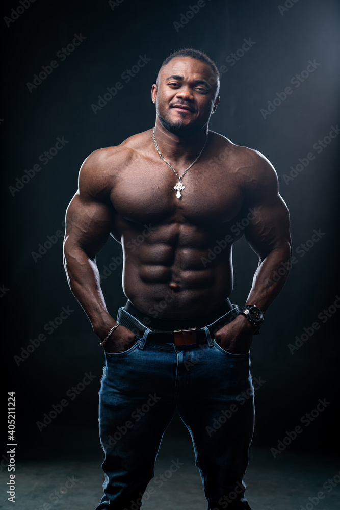 Muscular african american athlete with arms in pockets looking at camera. Black studio background.