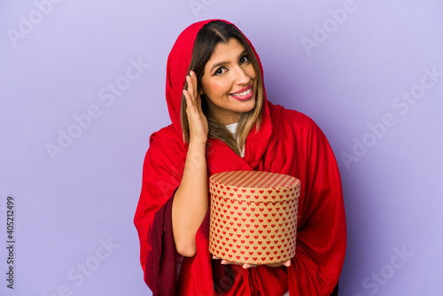 Young indian woman wearing a hijab holding a valentines day gift isolated trying to listening a gossip.