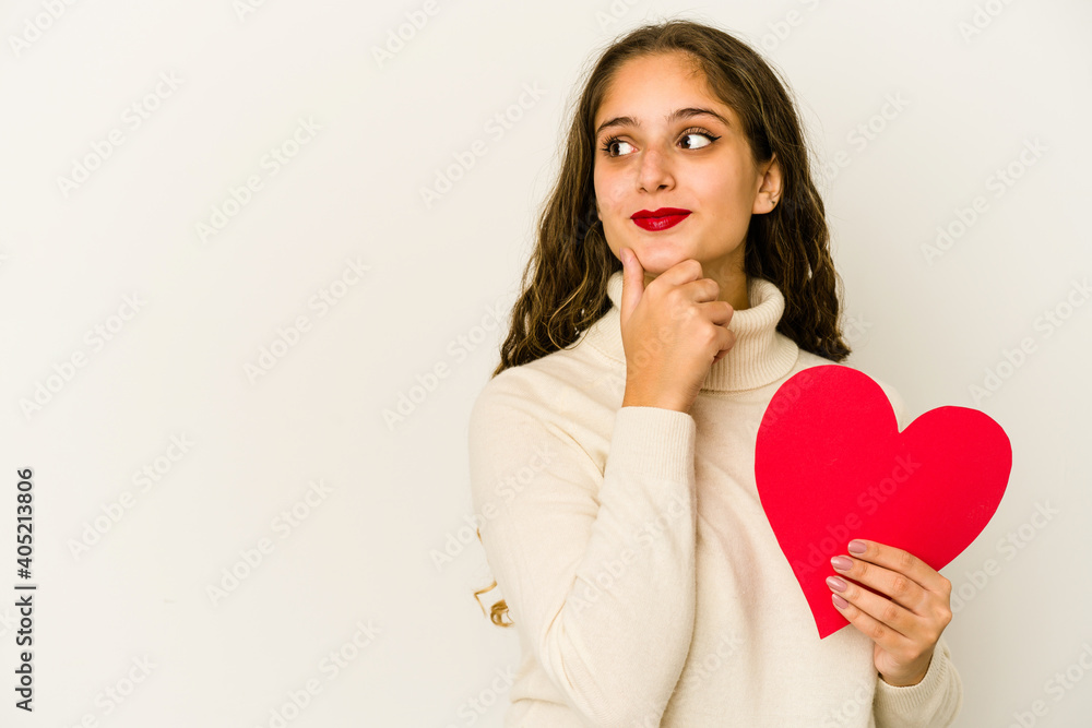 Young caucasian woman holding a heart valentines day shape isolated looking sideways with doubtful and skeptical expression. Young caucasian woman holding a heart valentines day shape isolated looking sideways with doubtful and skeptical expression.