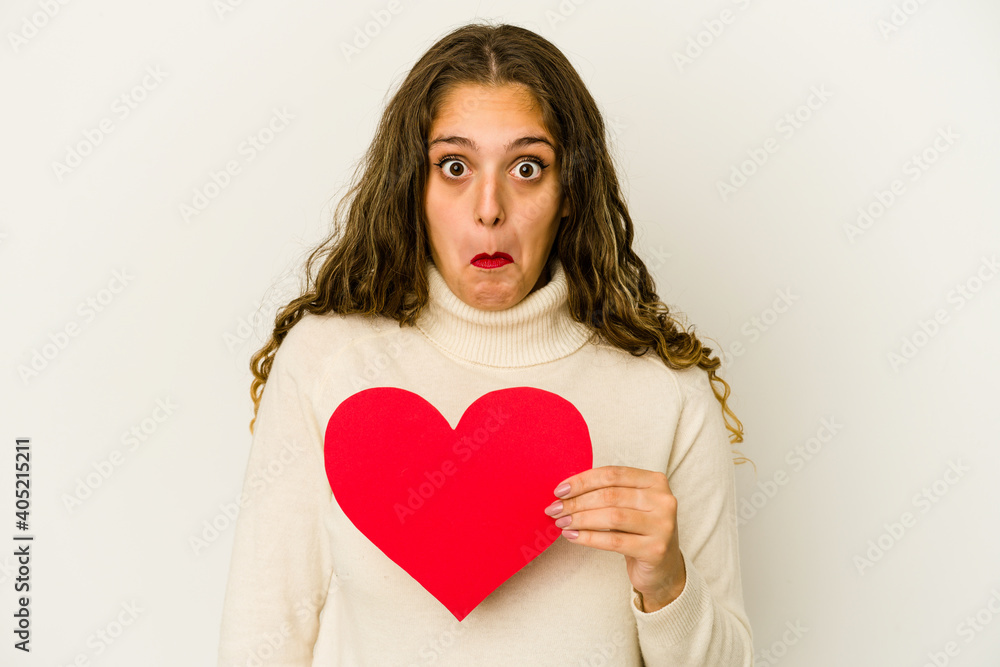 Young caucasian woman holding a heart valentines day shape isolated shrugs shoulders and open eyes confused. Young caucasian woman holding a heart valentines day shape isolated shrugs shoulders and open eyes confused.