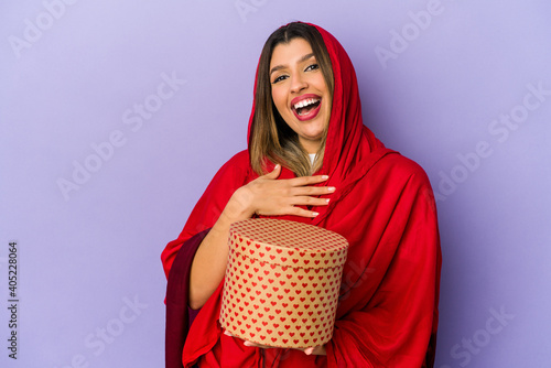 Young indian woman wearing a hijab holding a valentines day gift isolated laughs out loudly keeping hand on chest.