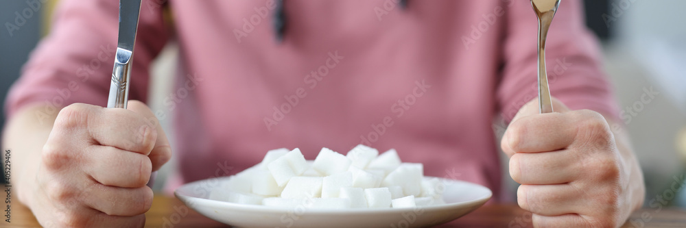 Woman holding fork and knife in hands near plate with lumps of sugar in kitchen. Unhealthy food concept Woman holding fork and knife in hands near plate with lumps of sugar in kitchen. Unhealthy food concept
