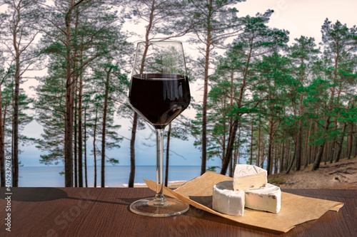 Glass of red wine with brie cheese against view of coniferous forest and blue sea
