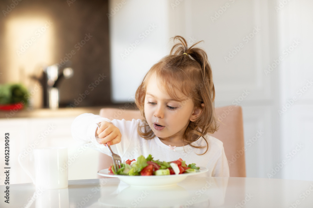Child girl eating fresh vegetables salad indoors sitting at kitchen table.Kid healthy food nutrition concept