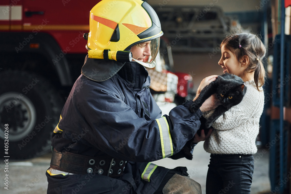 Cute black cat. Happy little girl is with male firefighter in protective uniform Cute black cat. Happy little girl is with male firefighter in protective uniform
