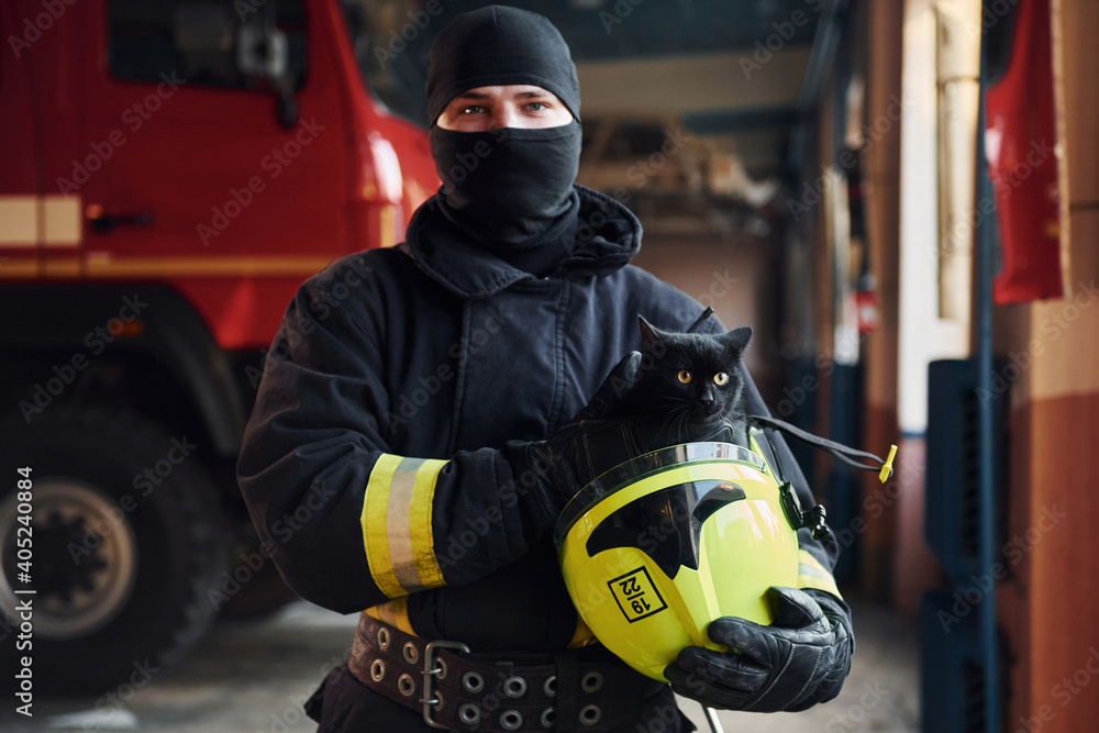 Naklejka premium Portrait of firefighter in protective uniform that holds cute little black cat