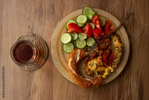 Selective focus, traditional Turkish tea in tea glass and Turkish bagel, tomato slices, cucumber slices in the plate on a wooden table. daylight comes through the window
