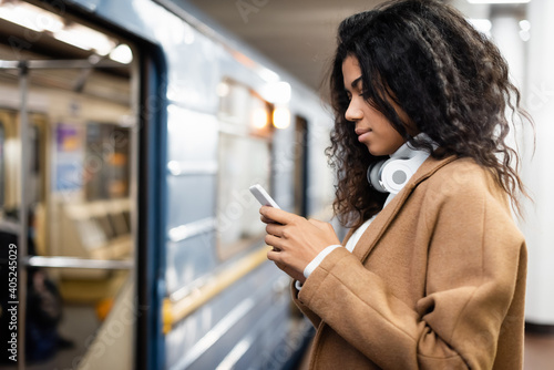 side view of african american woman in wireless headphones using smartphone in subway