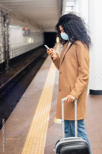 african american woman in medical mask standing with luggage and using smartphone in subway