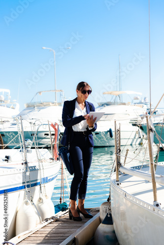 Female yachting dealer in marine blue suit holding notepad, standing outside a luxury sailboat, Heraklion, Crete, Greece