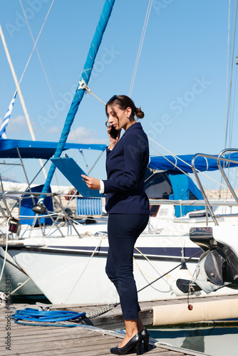 Female yachting dealer in marine blue suit holding notepad, standing outside a luxury sailboat using cell phone, Heraklion, Crete, Greece