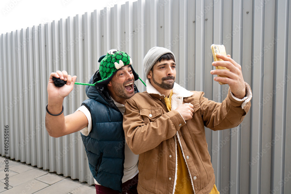 two friends greeting by video call with a yellow mobile smartphone with music with a funny hat super happy happy having a cool time with a metallic gray background in the underground street life style