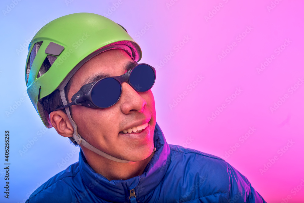 Young latin man smiling with green climbing helmet and blue jacket over colorful background Young latin man smiling with green climbing helmet and blue jacket over colorful background