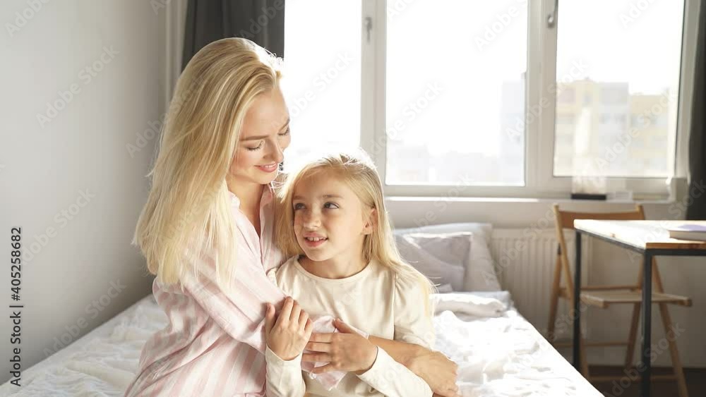 Portrait of friendly mother and child girl in bedroom looking at camera posing, hugging. Good relationships between parent and daughter. Portrait of friendly mother and child girl in bedroom looking at camera posing, hugging. Good relationships between parent and daughter.