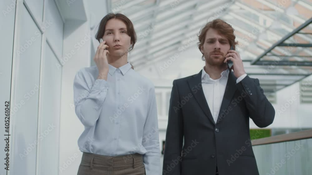 Medium shot of young Caucasian businesswoman and her handsome male coworker both wearing office wear going along business center hall and talking on telephones Medium shot of young Caucasian businesswoman and her handsome male coworker both wearing office wear going along business center hall and talking on telephones