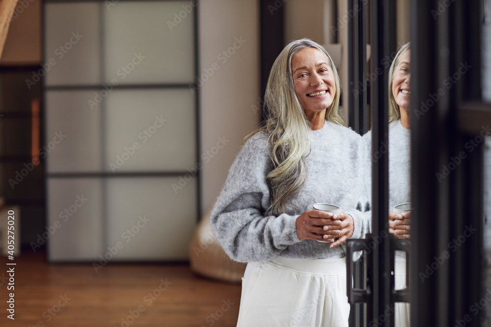 Smiling mature woman with gray hair holding coffee cup while leaning on door at home Smiling mature woman with gray hair holding coffee cup while leaning on door at home
