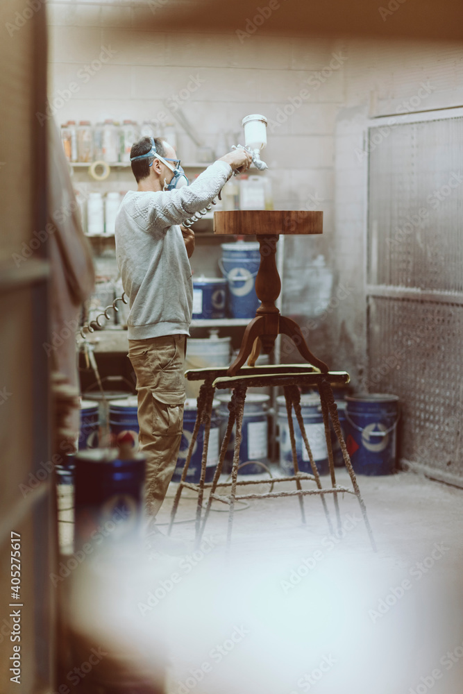 Manual worker with protective face mask lacquering table while standing at workshop
