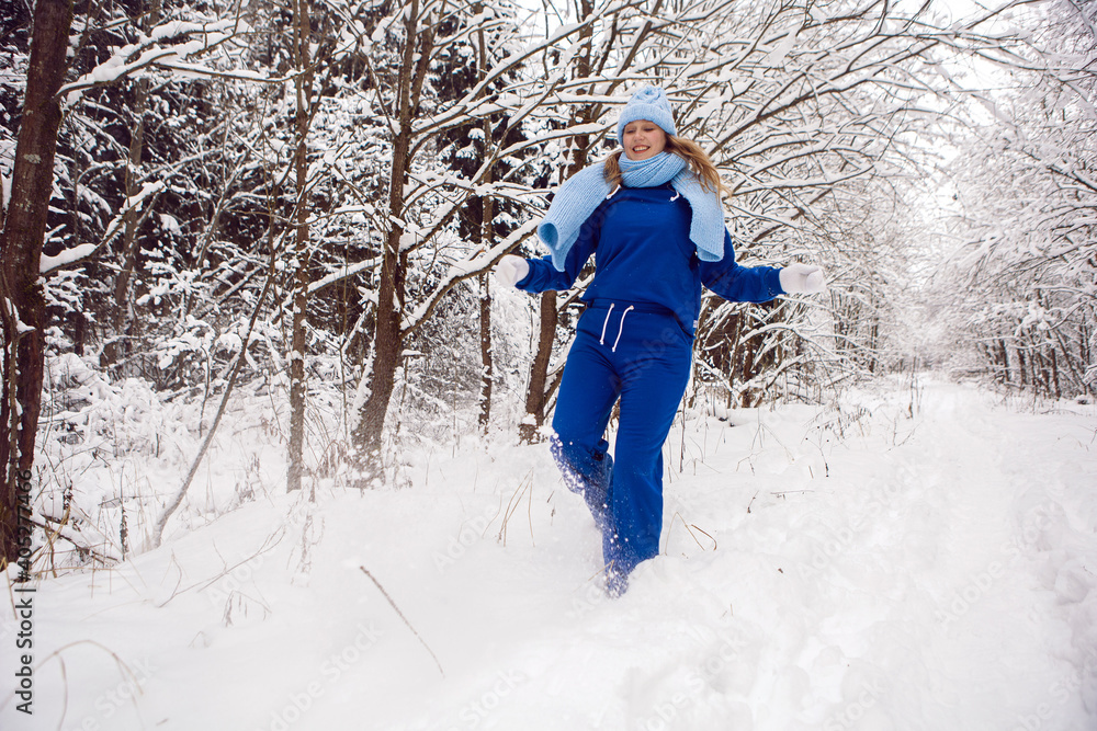 woman in a blue tracksuit white mittens and scarf stands in winter in a snow covered forest