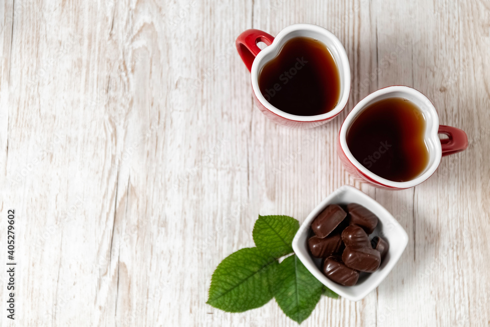 Two red cups with tea on a wooden background. Breakfast for lovers. Heart shaped cup. Valentine's Day. Chocolates in a vase. View from above. Two red cups with tea on a wooden background. Breakfast for lovers. Heart shaped cup. Valentine's Day. Chocolates in a vase. View from above.