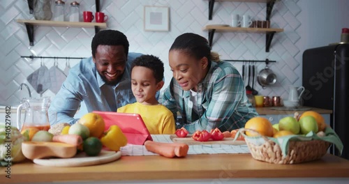Candid african american couple aof parents with son calling on tablet computer video chat conference rejoicing family dinner standing in the kitchen. photo