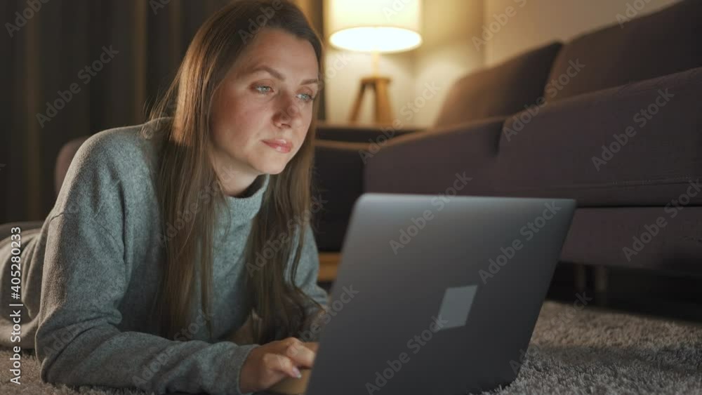 Woman is lying on the carpet and working on a laptop in the evening. Concept of remote work. Woman is lying on the carpet and working on a laptop in the evening. Concept of remote work.