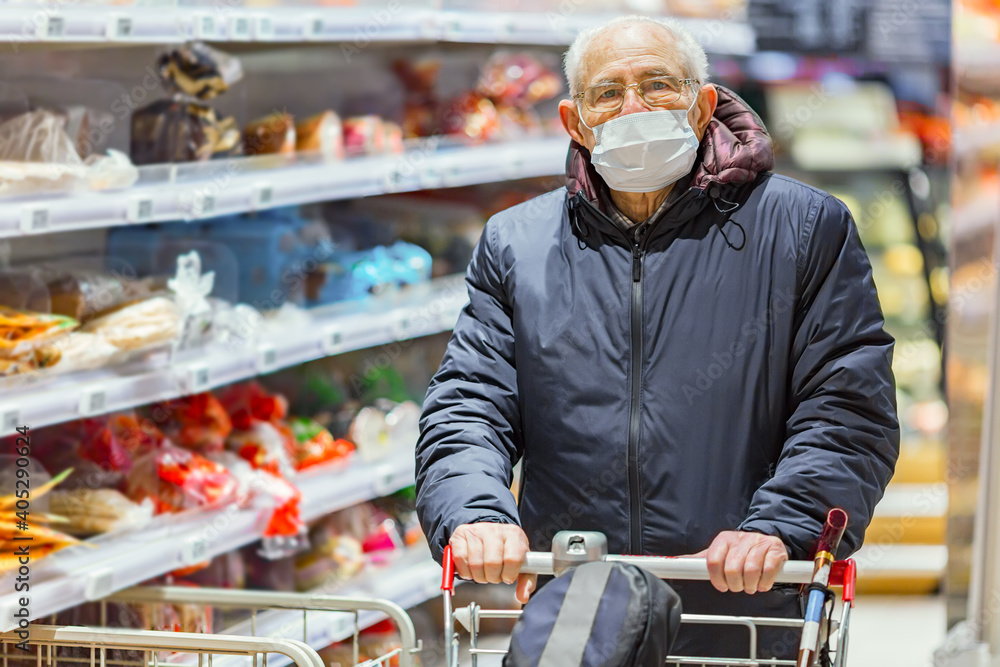 Old senior european man wearing protective facial mask pushing shopping cart in the supermarket. Shopping during COVID-19 concept.