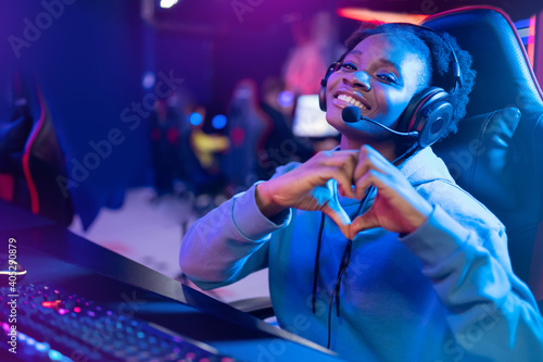 Streamer African beautiful girl shows heart sign with hands professional gamer playing online games computer, neon color photo