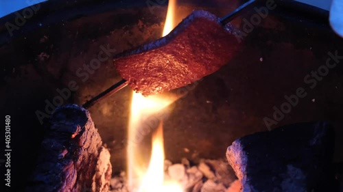 Outdoor picnic with a fire pit. Traditional Turkish sausage, SUJUK, on a metallic shish skewer is hanging on a wooden fire. The orange flame is frizzling the meat and fat is boiling. photo