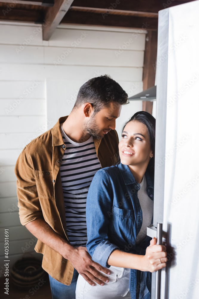 smiling woman opening fridge and looking at man seducing her in kitchen smiling woman opening fridge and looking at man seducing her in kitchen