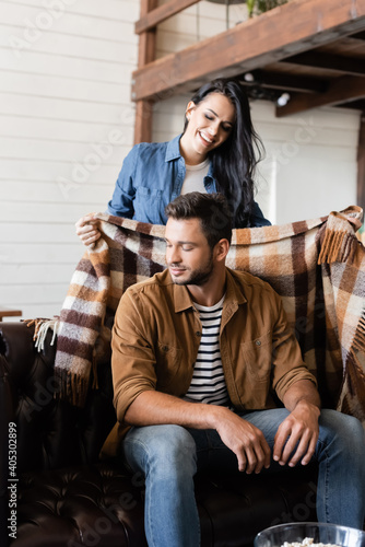 happy woman covering boyfriend with warm plaid blanket at home
