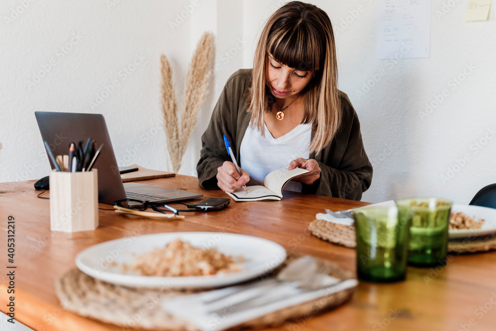 Female professional with food plate on table writing on notepad in home office Female professional with food plate on table writing on notepad in home office