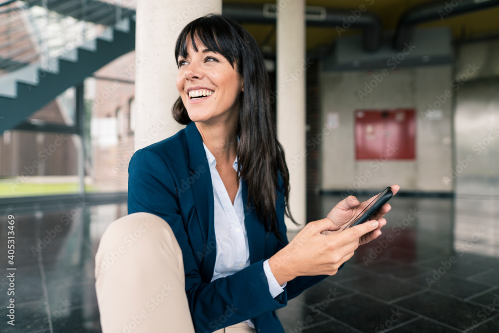Smiling businesswoman looking away while holding smart phone in office Smiling businesswoman looking away while holding smart phone in office