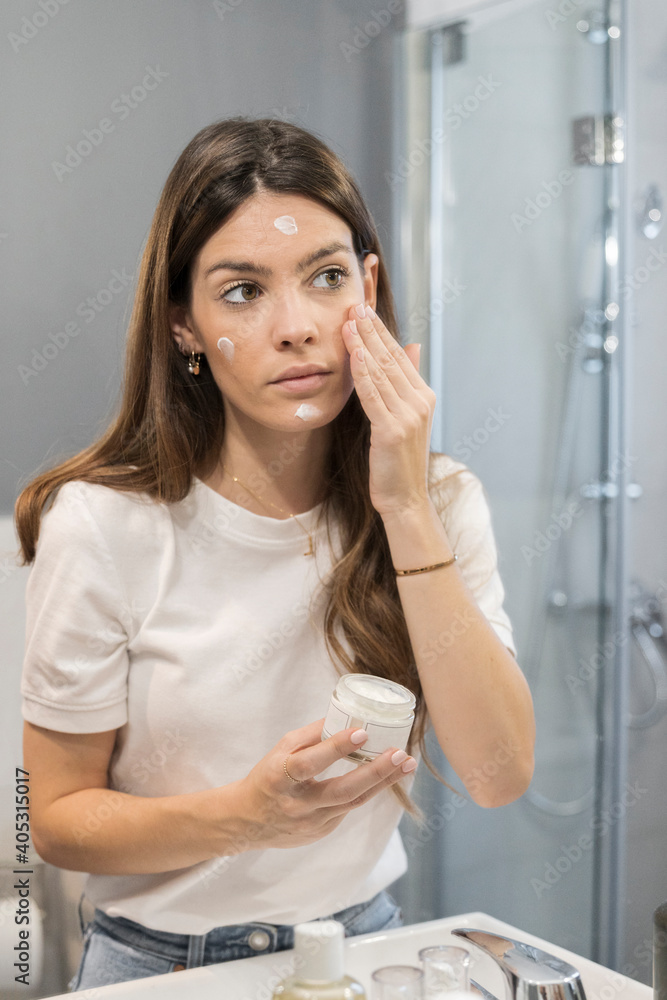 Young woman rubbing cream on face while standing in bathroom at home Young woman rubbing cream on face while standing in bathroom at home