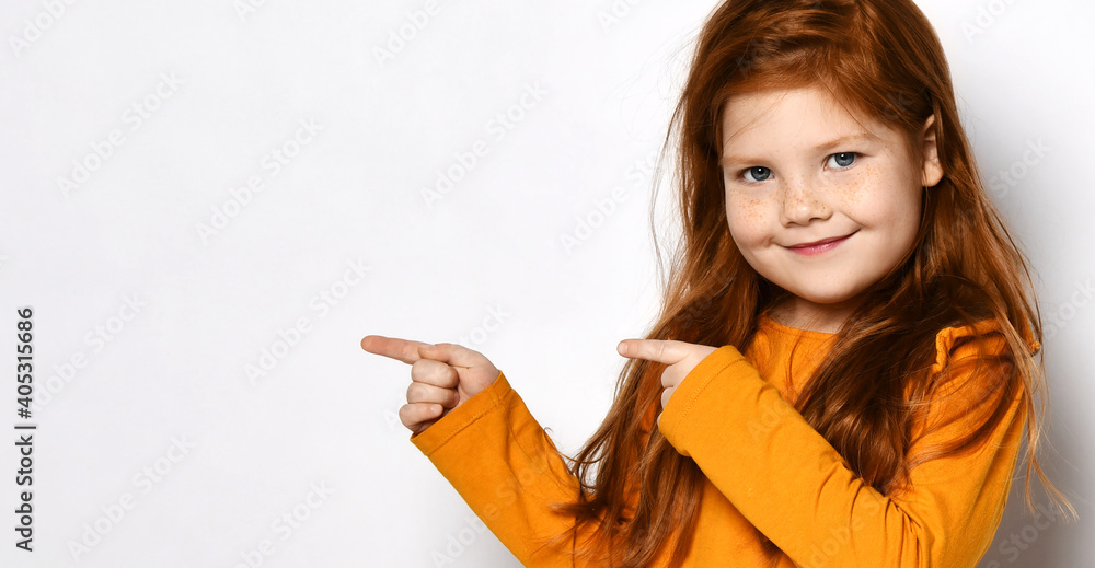 Smiling small red haired girl with freckles in yellow comfortable longsleeve standing and pointing aside with fingers over white background, copy space. Happy childhood, stylish look concept Smiling small red haired girl with freckles in yellow comfortable longsleeve standing and pointing aside with fingers over white background, copy space. Happy childhood, stylish look concept