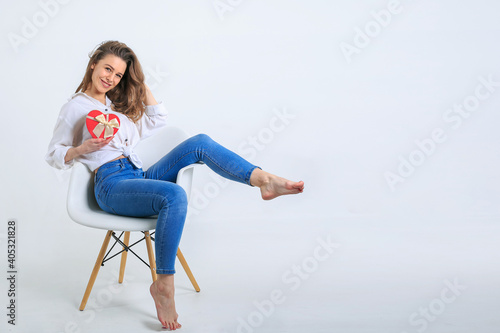 Beautiful cheerful happy young brunette woman with long curly hair in white shirt and jeans sitting on white chair and holds red gift heart-shaped with ribbon isolated on white. March 8th Holiday Conc