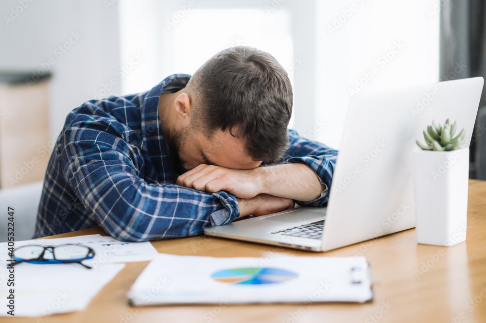Obraz premium Tired freelancer man is fall asleep at the workplace. Exhausted busy guy working overtime, sits at the desk at home, need a rest, overwork concept. Work from home